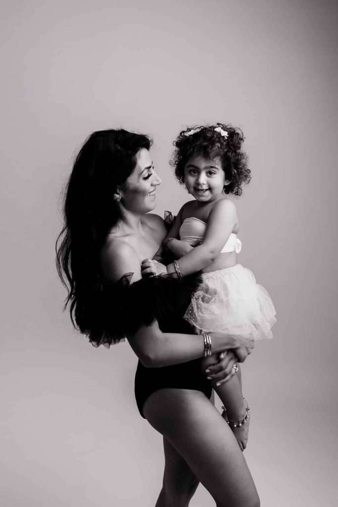 Woman in a black off-shoulder outfit holds a smiling young girl in a tutu. Both are looking at each other in a studio with a plain background. The image is black and white.