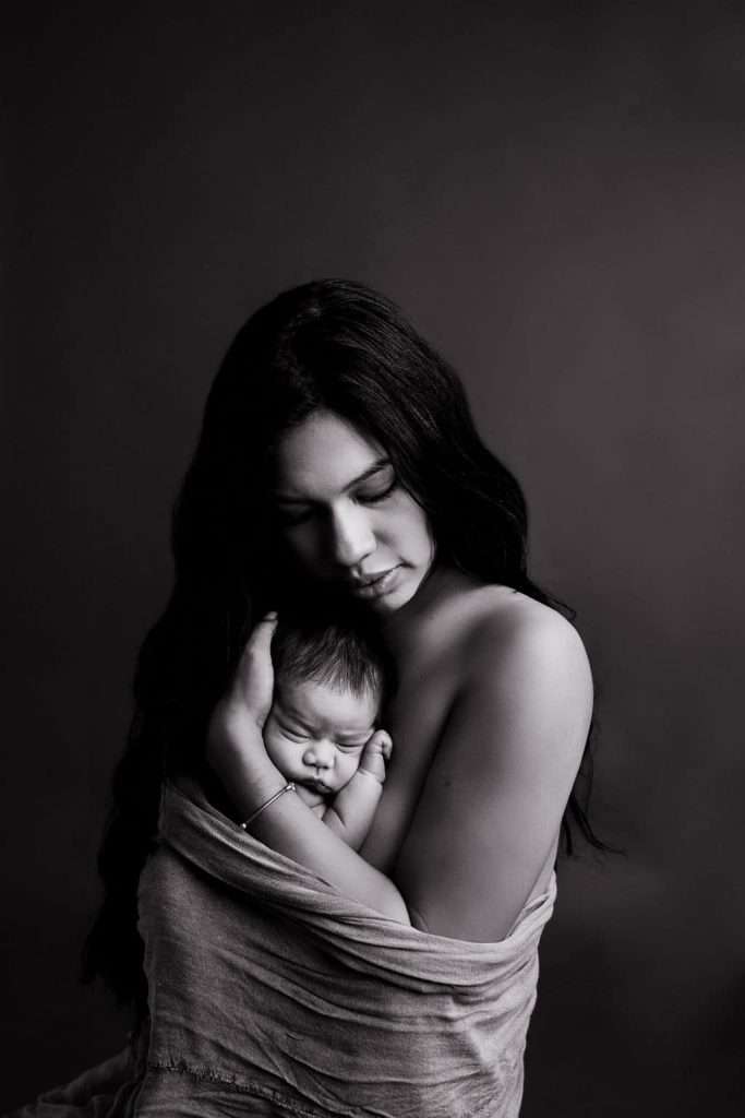 A woman with long dark hair holds a sleeping newborn close to her chest. The photo is in black and white with a plain background.