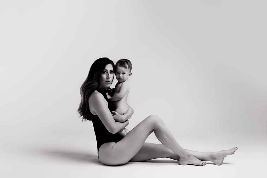 A woman in a black bodysuit sits on the floor holding a naked baby, both looking at the camera, against a plain light background.