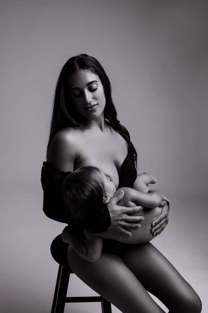 A woman seated on a stool breastfeeds a baby, holding the child in her arms. The image is in black and white.