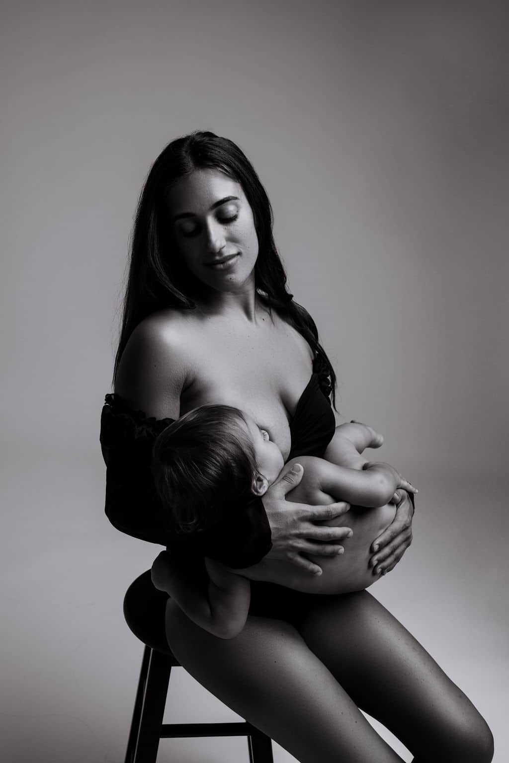 A woman seated on a stool breastfeeds a baby, holding the child in her arms. The image is in black and white.