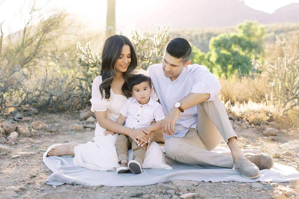A family of three sits on a blanket outdoors in a desert landscape, with the parents on either side of their young child, all dressed in light-colored clothing.