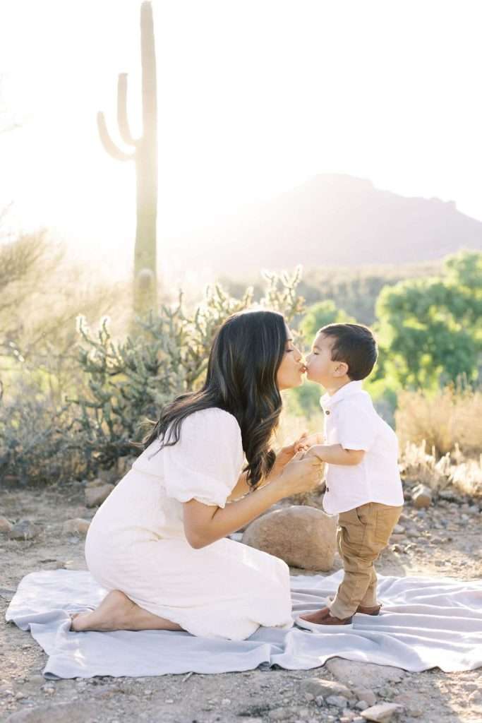 Woman in a white dress kneels on a blanket outdoors, gently touching noses with a young boy. Desert landscape with cacti and mountains in the background.