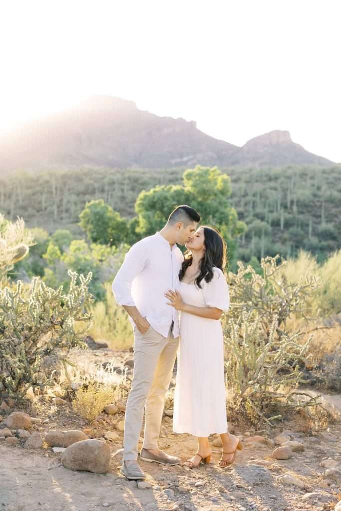 A couple stands in a desert landscape at sunset, the man kissing the woman’s forehead while they both wear light-colored clothing. Shrubs and mountains are visible in the background.