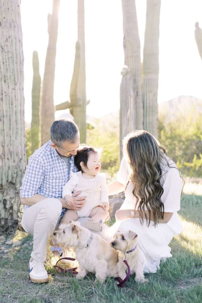 A man and woman kneel beside a crying baby and two small dogs in front of tall cacti in a sunlit outdoor setting.