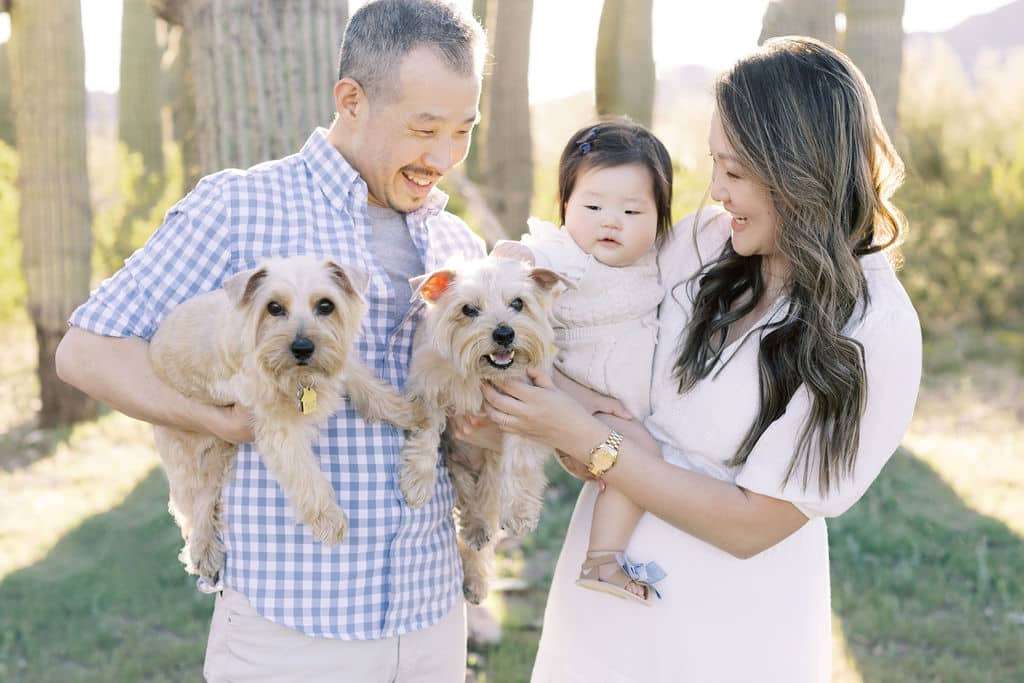 Two adults hold a baby and two small dogs outdoors on a sunny day, standing in front of tall cacti and greenery.