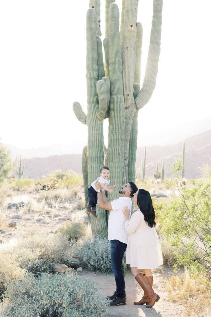 A man lifts a small child while a woman stands nearby in a desert landscape with large cacti and mountains in the background.