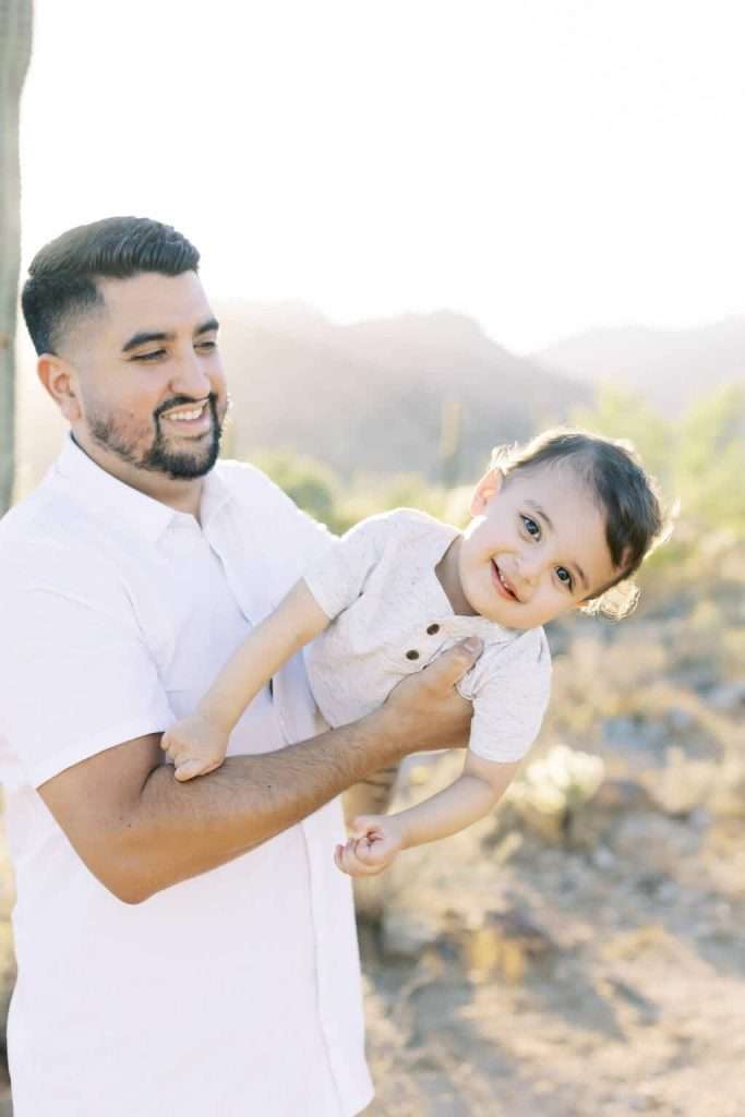 A man in a white shirt holds a smiling young child sideways outdoors with sunlight and desert landscape in the background.