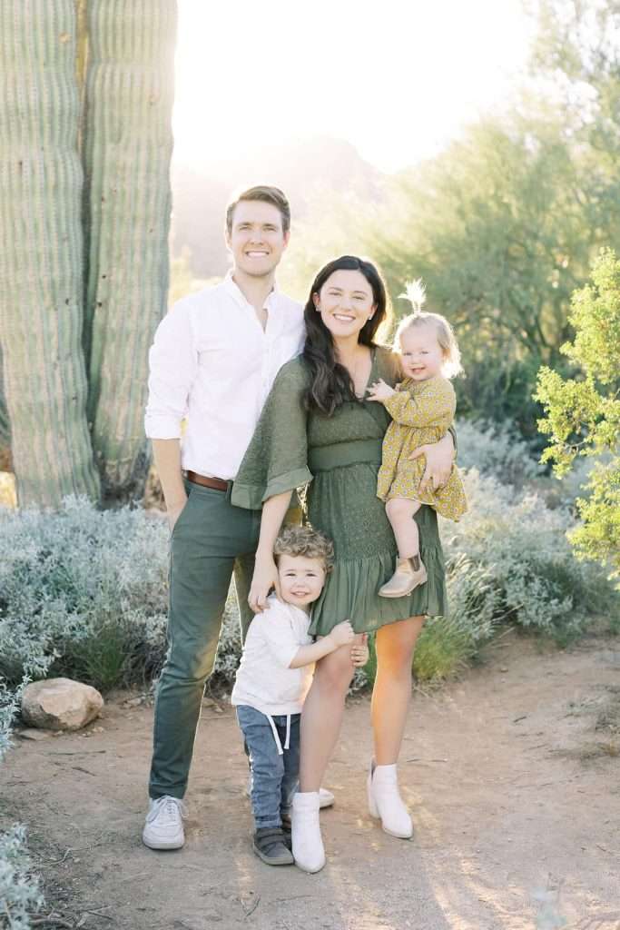 A family of four poses outdoors near a large cactus, with parents standing and two young children, one held by the mother, all smiling at the camera in natural light.