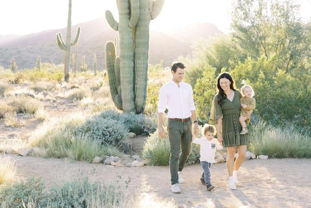 A family of four walks on a desert trail with cacti and shrubs, holding hands. The parents are dressed in green and white, and the sun is shining in the background.