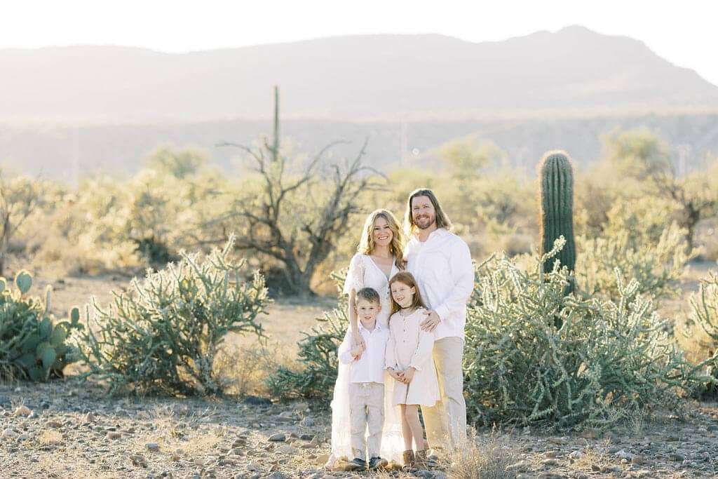 A family of four stands together in a desert landscape with cacti and mountains in the background, all wearing light-colored clothing.