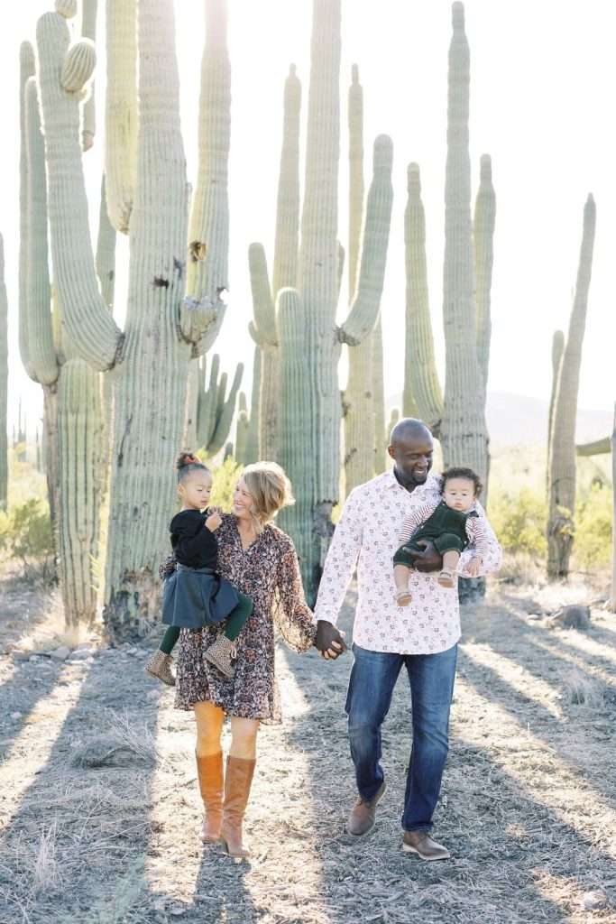 A family of four walks in a desert landscape with tall cacti. The parents each carry a child and hold hands, all dressed in casual outfits, sunlight shining through the cacti.