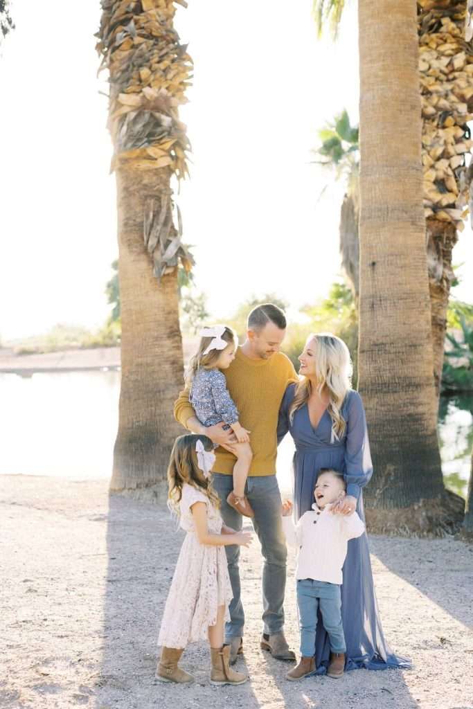 A family of five stands outdoors between palm trees, smiling and interacting with each other on a sunny day.