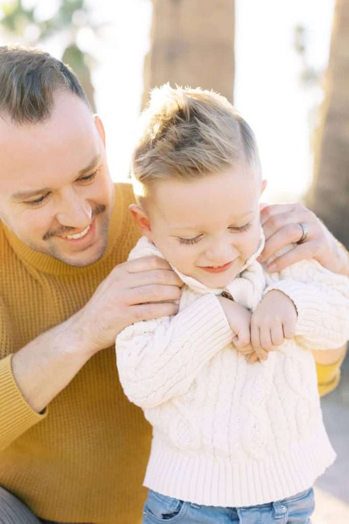 An adult kneels beside a young child outdoors, gently holding the child’s shoulders. Both are wearing sweaters and appear to be enjoying a sunny day.