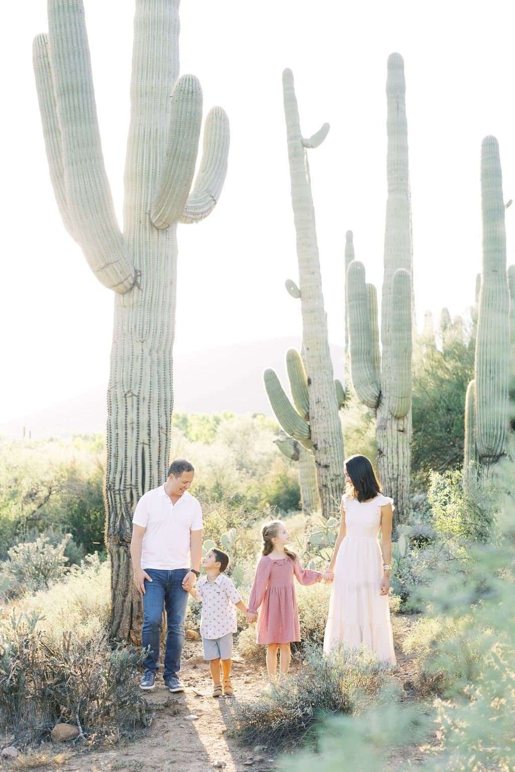 A family of four stands holding hands on a desert path surrounded by tall cacti and greenery, with sunlight streaming in the background.