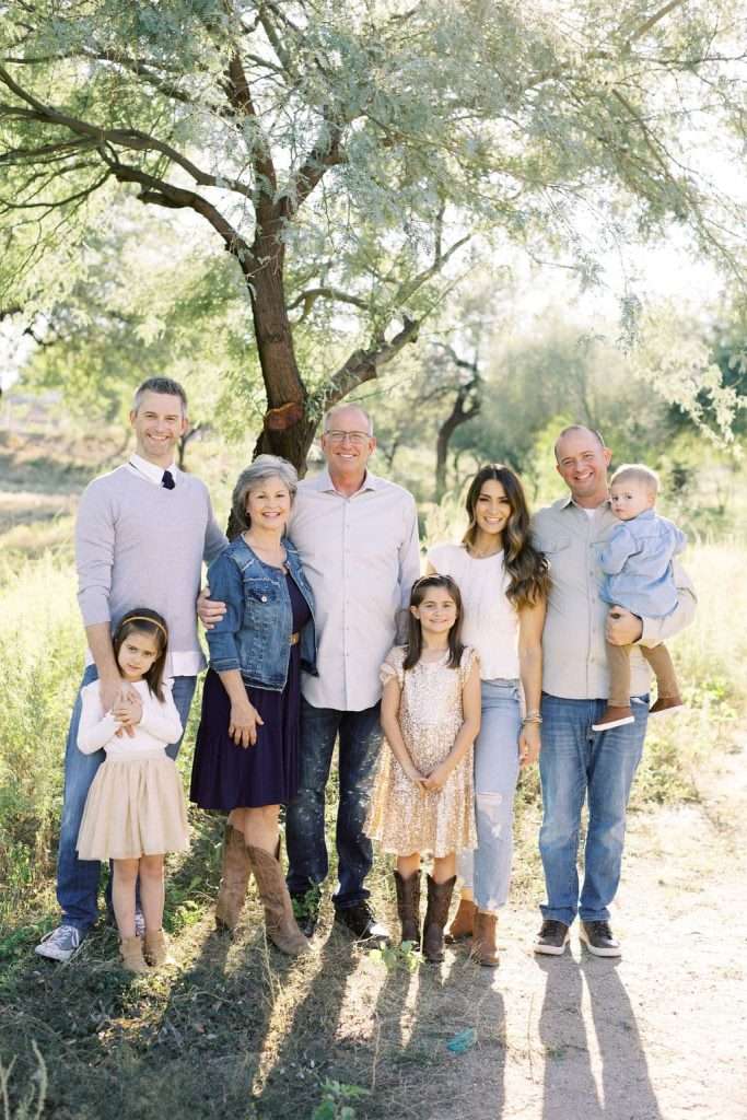 Three men, two women, two girls, and a toddler stand together outdoors in front of a tree on a sunny day, smiling at the camera.