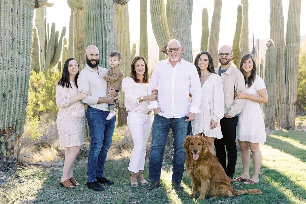 Eight adults, one child, and a golden retriever pose together in front of large cacti outdoors on a sunny day.