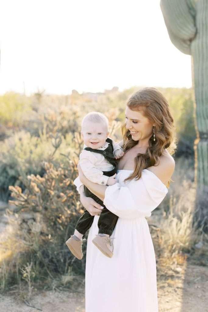 Woman in a white dress holds a smiling baby outdoors in a desert landscape with cacti and shrubs in the background.