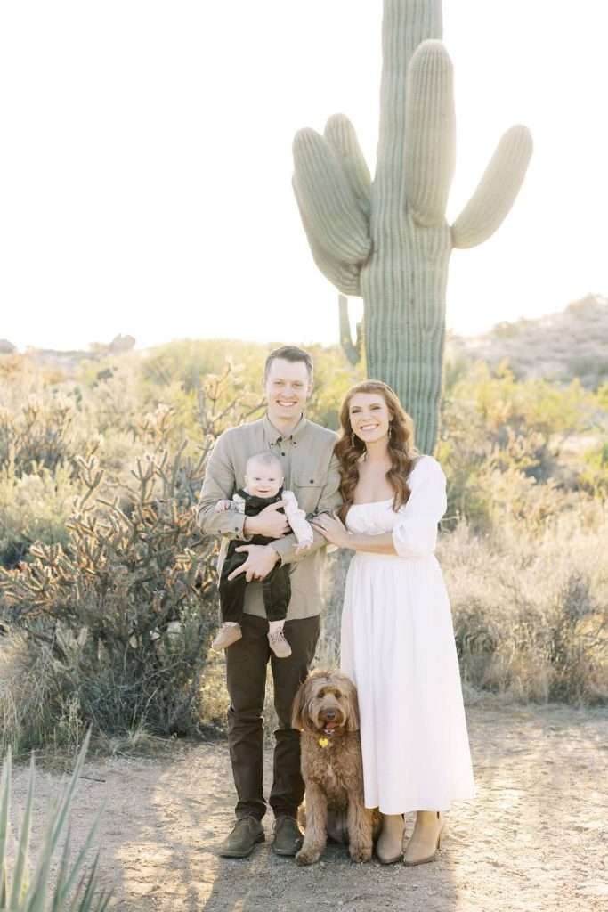 A man, woman, baby, and brown dog pose together outdoors in front of a large cactus in the desert, all smiling at the camera.