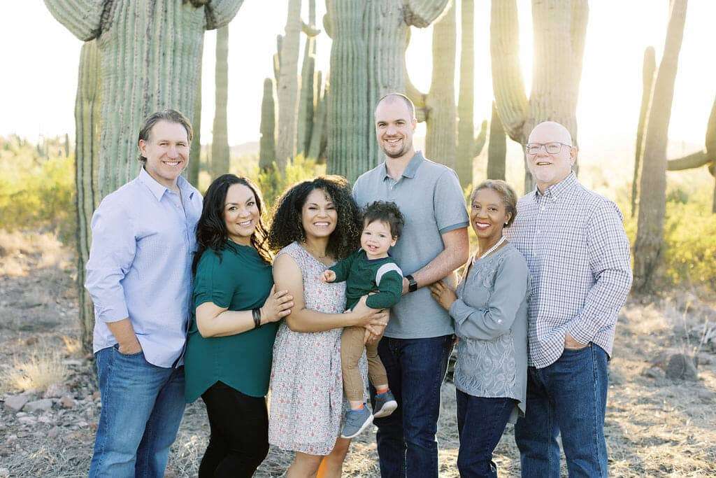 Seven people, including a child, pose together outdoors in front of tall cacti, all smiling at the camera in casual clothing.