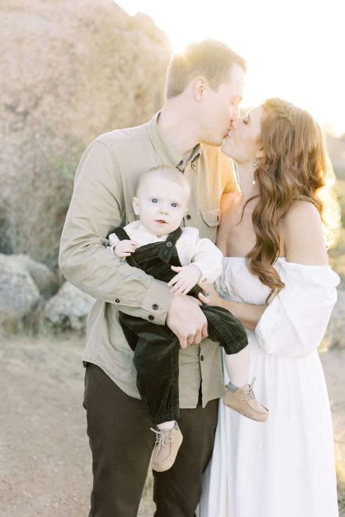 A man and woman kiss while the man holds a baby in his arms. They stand outdoors in sunlight, with rocks in the background.