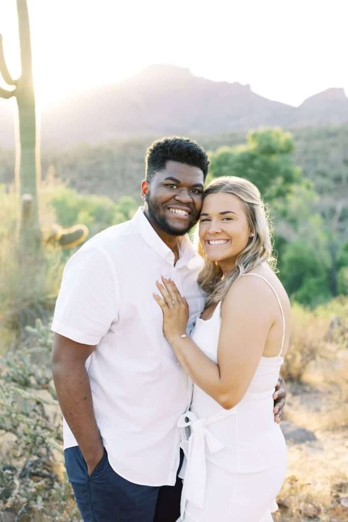A couple stands close together outdoors, smiling at the camera with desert plants and mountains in the background.