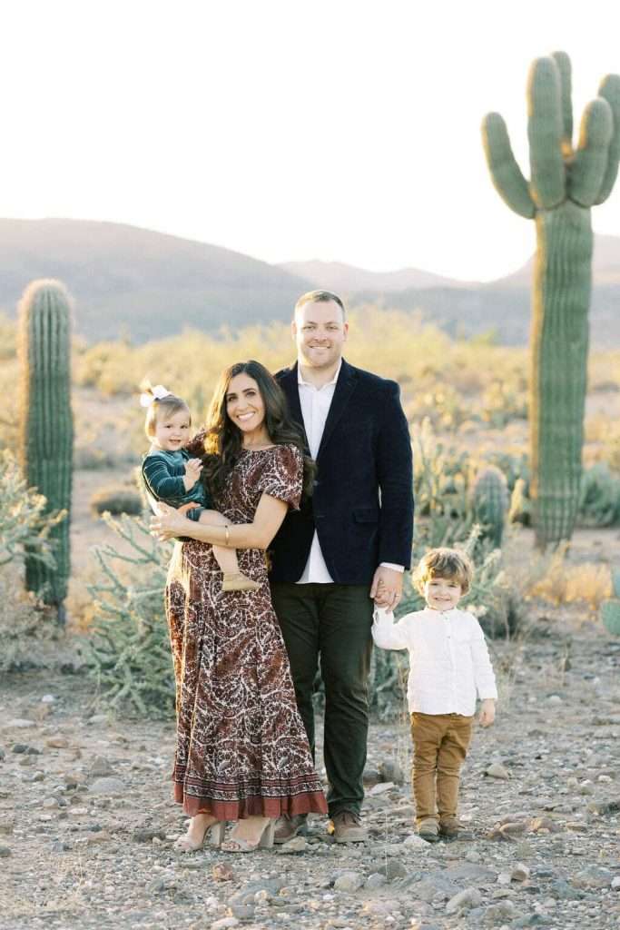 A family of four poses together in a desert landscape with cacti, parents standing close with their young son and daughter, all dressed in coordinated outfits.