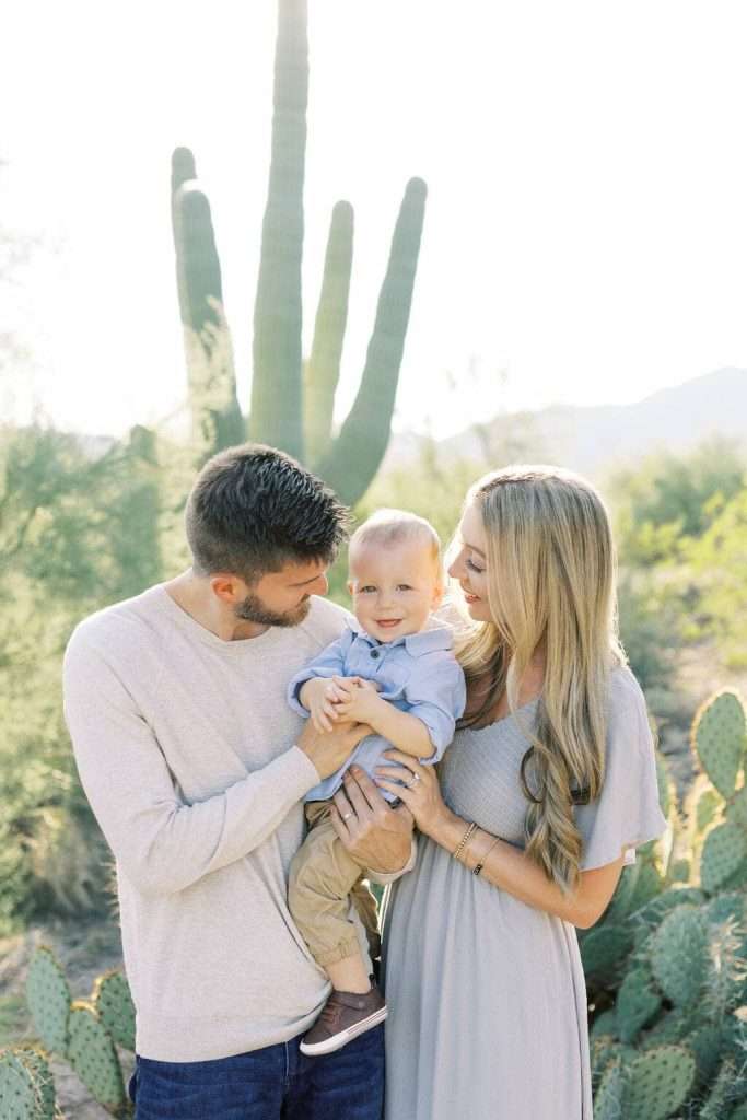 A man and woman hold a baby while standing outdoors in front of a tall cactus, with greenery and mountains in the background.