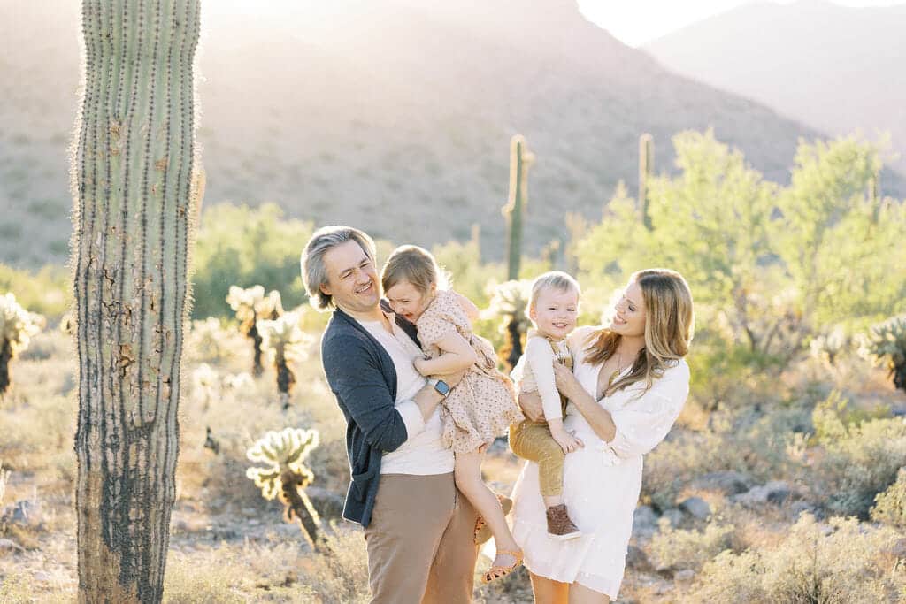 A family of four stands together in a desert landscape with cacti, smiling and laughing in sunlight.