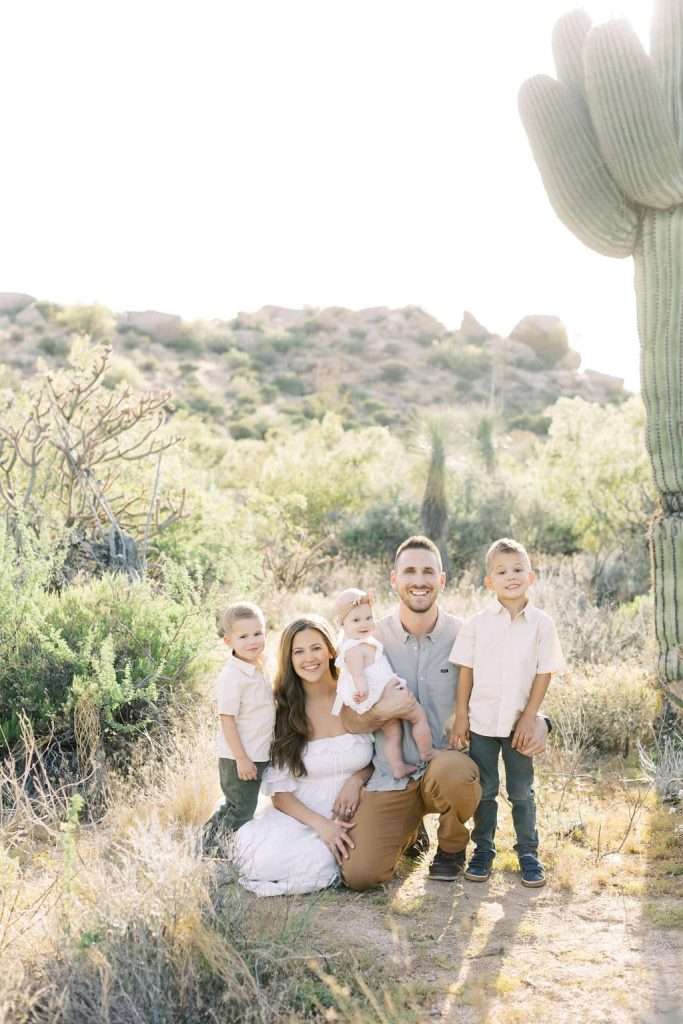 A family of five poses in a desert landscape near a cactus, with the parents and three young children smiling at the camera in sunlight.