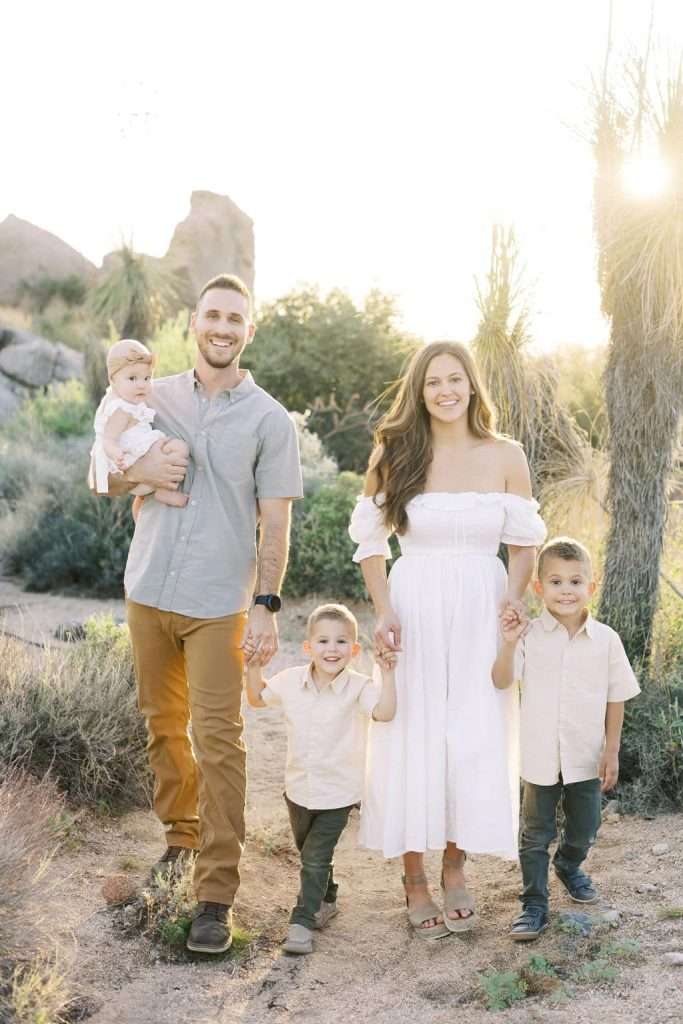A family of five stands outdoors in the sunlight, with a man holding a baby, a woman in a white dress, and two young boys holding hands.
