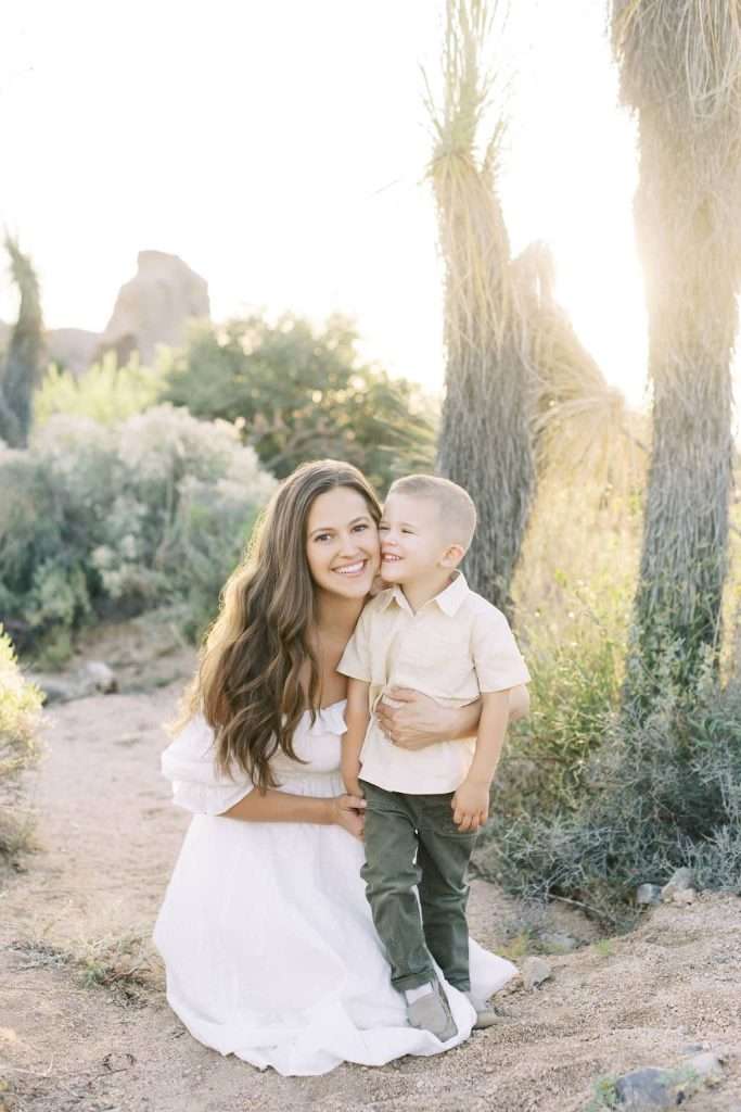Woman in a white dress kneels beside a young boy in light clothing, both smiling, with desert plants and sunlight in the background.