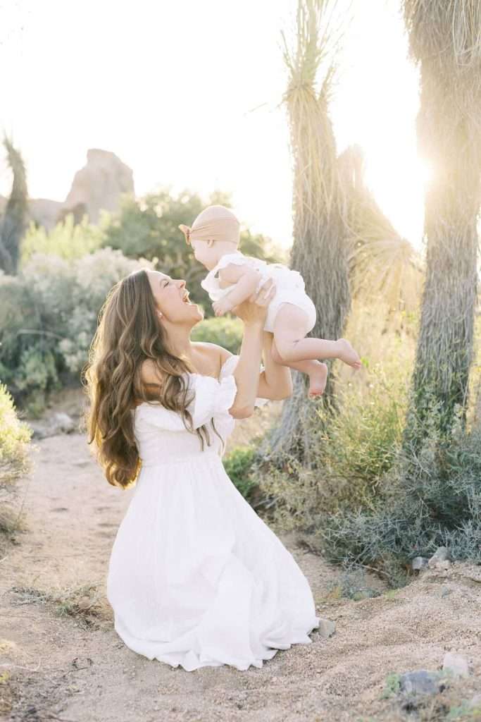 A woman in a white dress kneels on a sandy path, lifting a baby in white clothing, with desert plants and sunlight in the background.