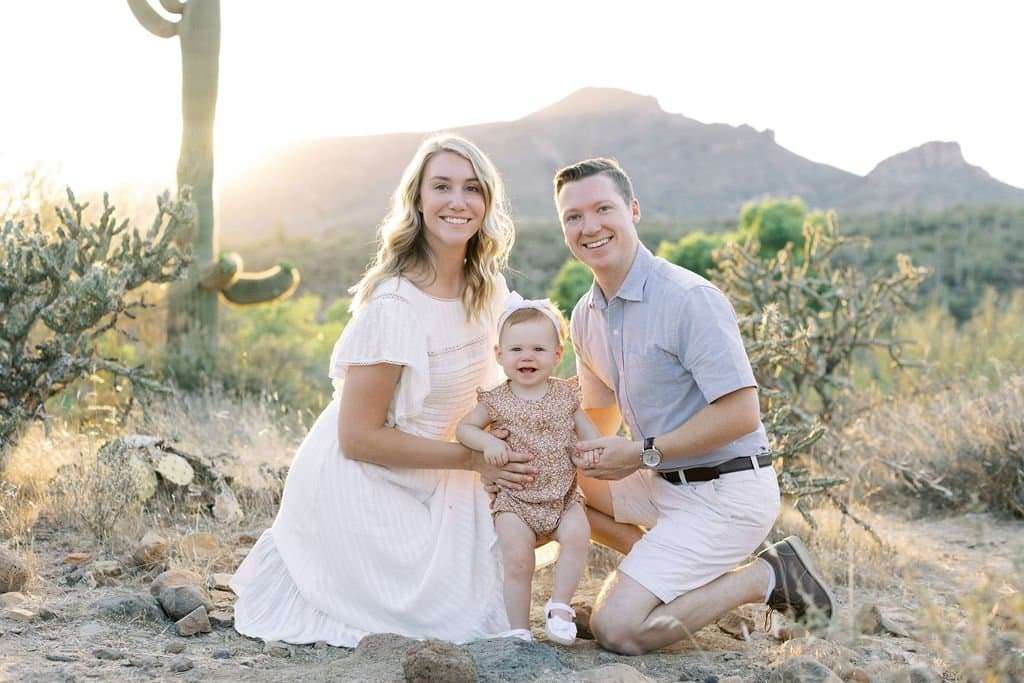 A woman, a man, and a young child pose together outdoors in a desert landscape with mountains and sunlight in the background.