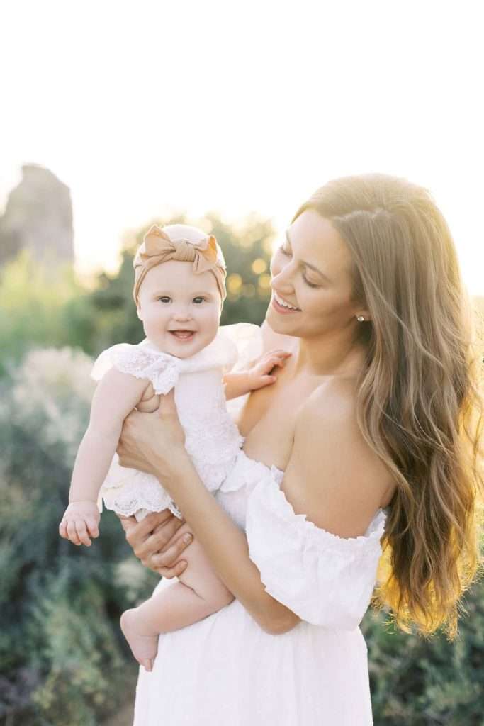 A woman with long hair in a white dress holds a smiling baby wearing a bow headband and white outfit outdoors in sunlight.