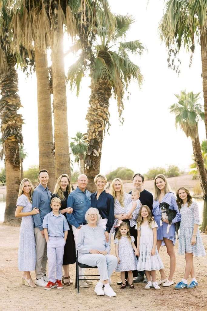 A multigenerational family poses outdoors near palm trees, with adults standing and children gathered around an elderly couple seated at the center.