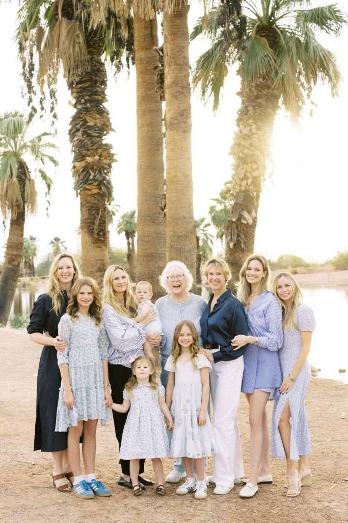 A group of eleven women and girls, including a baby, stand together outdoors in front of palm trees, smiling at the camera.