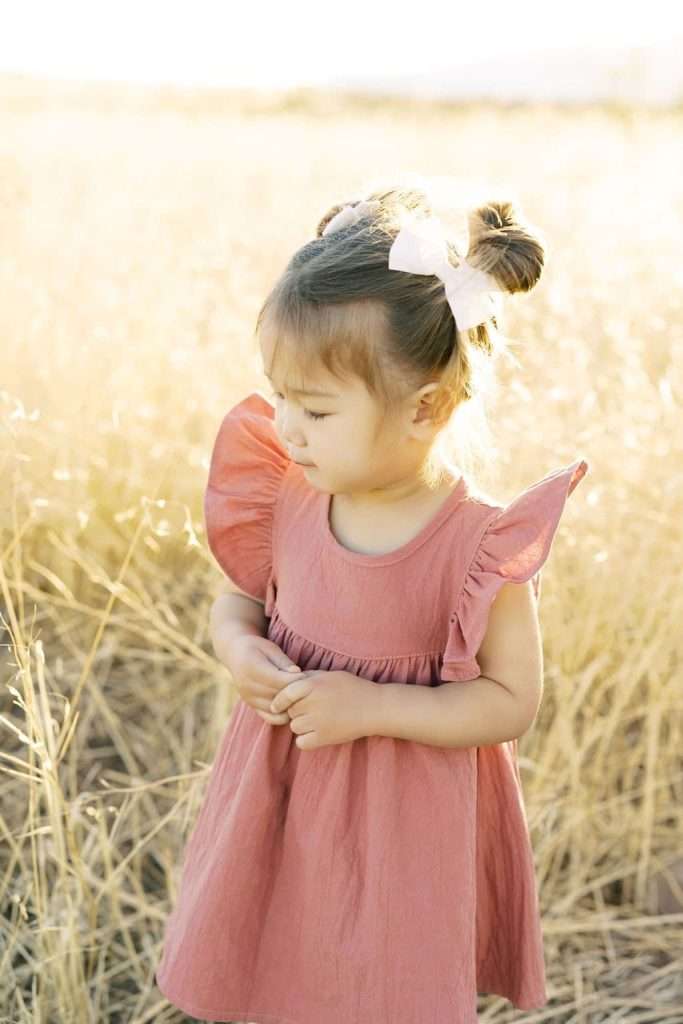 Young girl with pigtails and a white bow wears a pink dress and stands in a sunlit field of tall dry grass, looking down.