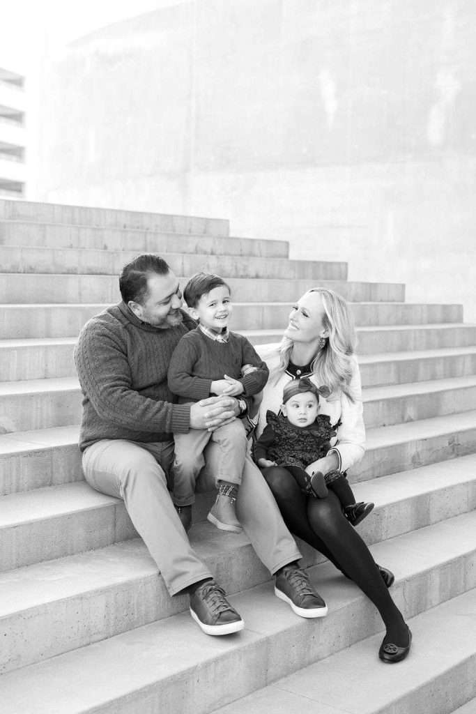 A man, woman, young boy, and baby sit together on outdoor steps, smiling and interacting with each other. The photo is in black and white.