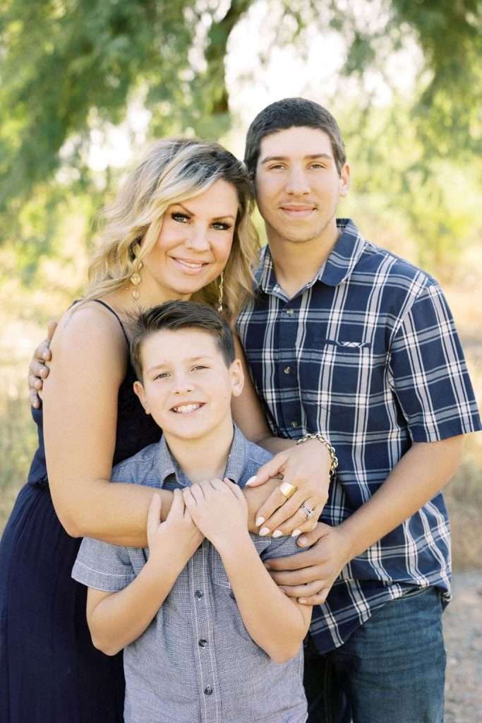 Woman with two boys poses outdoors, all smiling at the camera. They are dressed in casual shirts, standing closely together in front of a tree.