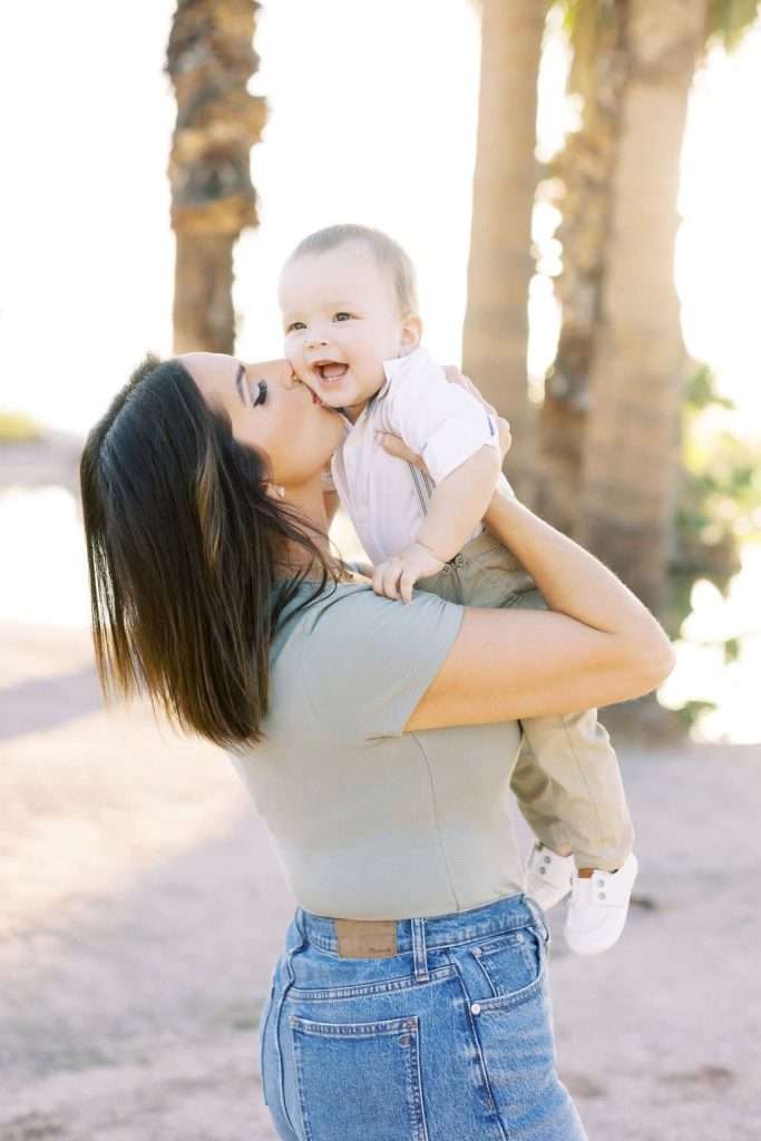 Woman holding and kissing a smiling baby outdoors with palm trees and sunlight in the background.