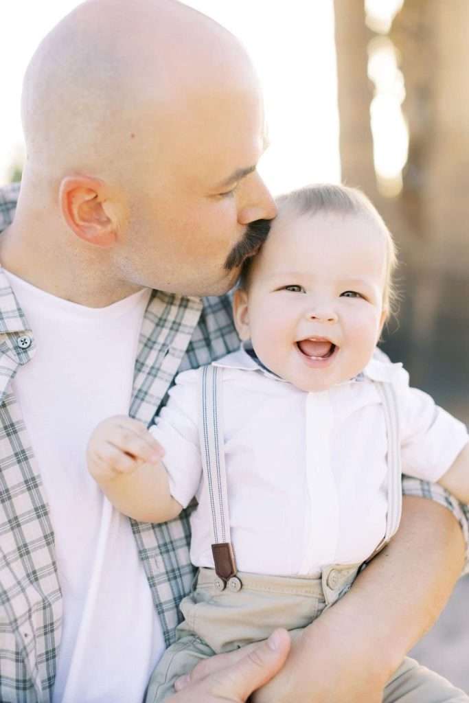 A man with a mustache kisses a smiling baby wearing suspenders while holding them in his arms outdoors.