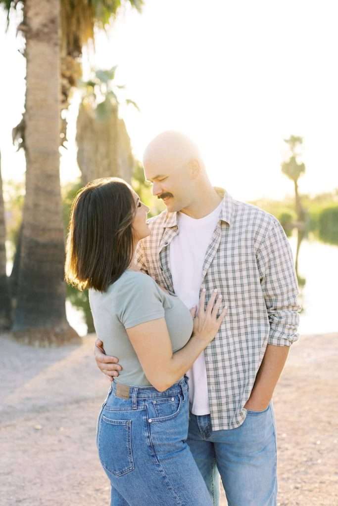 A couple stands close together outdoors by palm trees, facing each other and smiling, with sunlight shining behind them.
