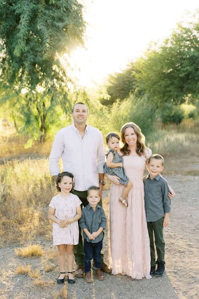 A family of six stands outdoors on a sunny day, with two adults and four children posing and smiling in front of green trees and dry grass.