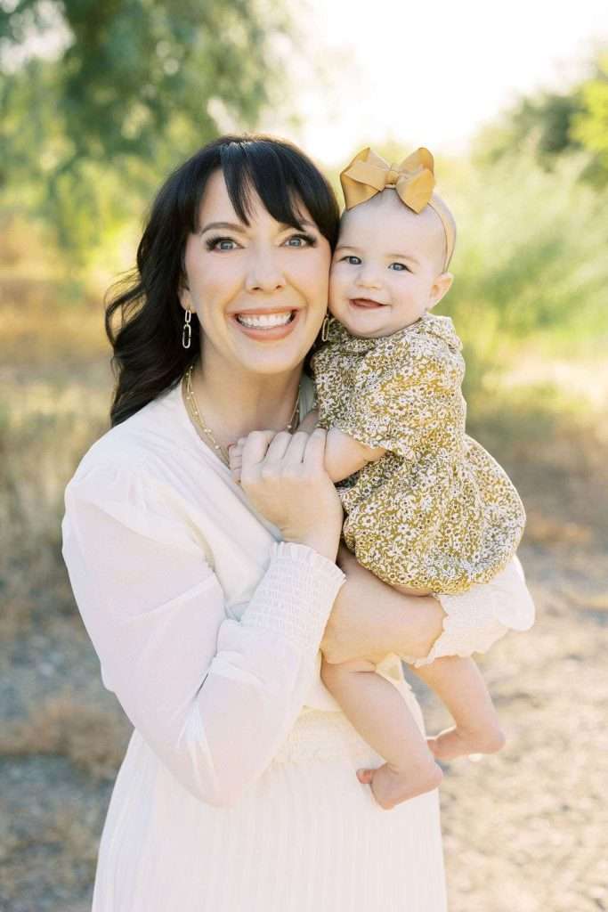 Woman in a white dress holds a smiling baby wearing a gold floral outfit and matching bow, standing outdoors with trees and sunlight in the background.