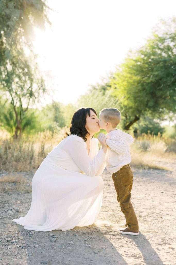 Woman in a white dress kneels outdoors to kiss a young boy holding her hands, with trees and sunlight in the background.