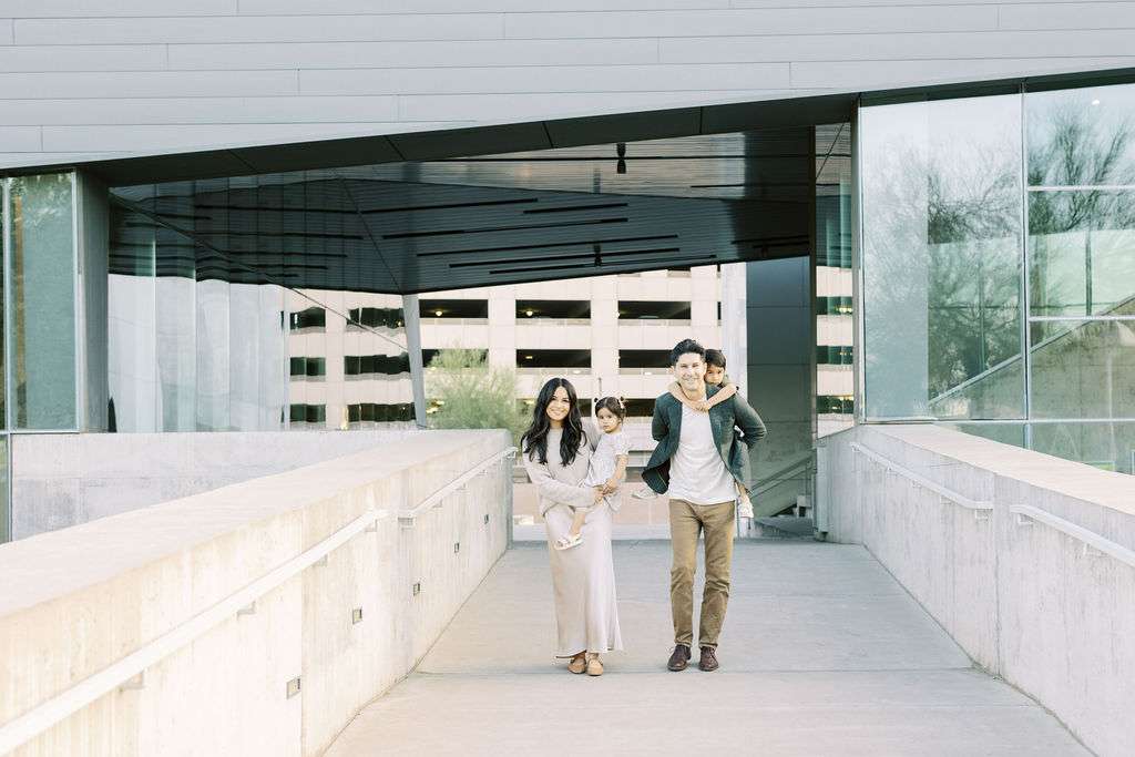 A family of four walks on a concrete pathway outside a modern building; two adults and two children, one child is carried and the other is on an adult’s shoulders.
