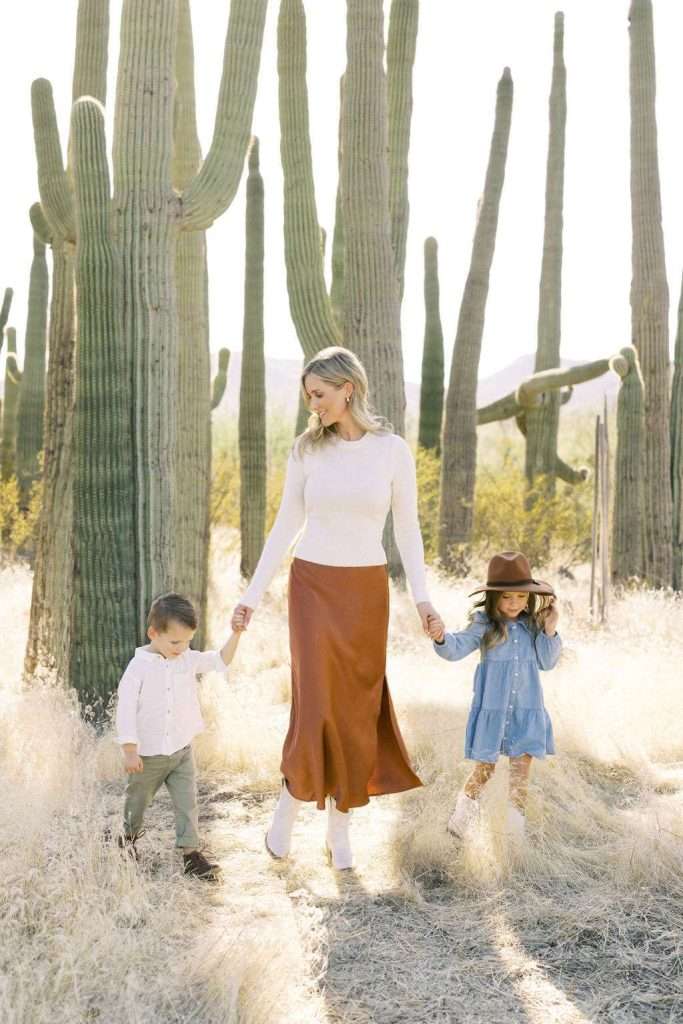 Woman in a white top and brown skirt holds hands with a young boy and girl, walking in a desert landscape with tall cacti and dry grass.