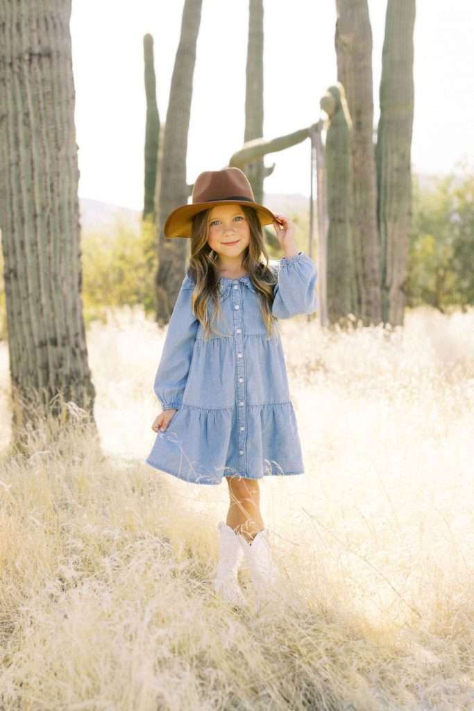 A young girl in a blue dress and brown hat stands in a field of tall dry grass with large cacti in the background.