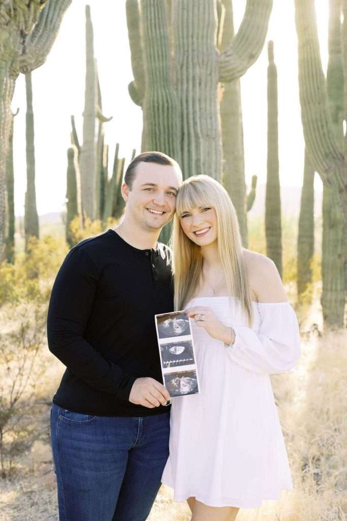 A smiling couple stands in a desert with cacti, holding ultrasound photos together.